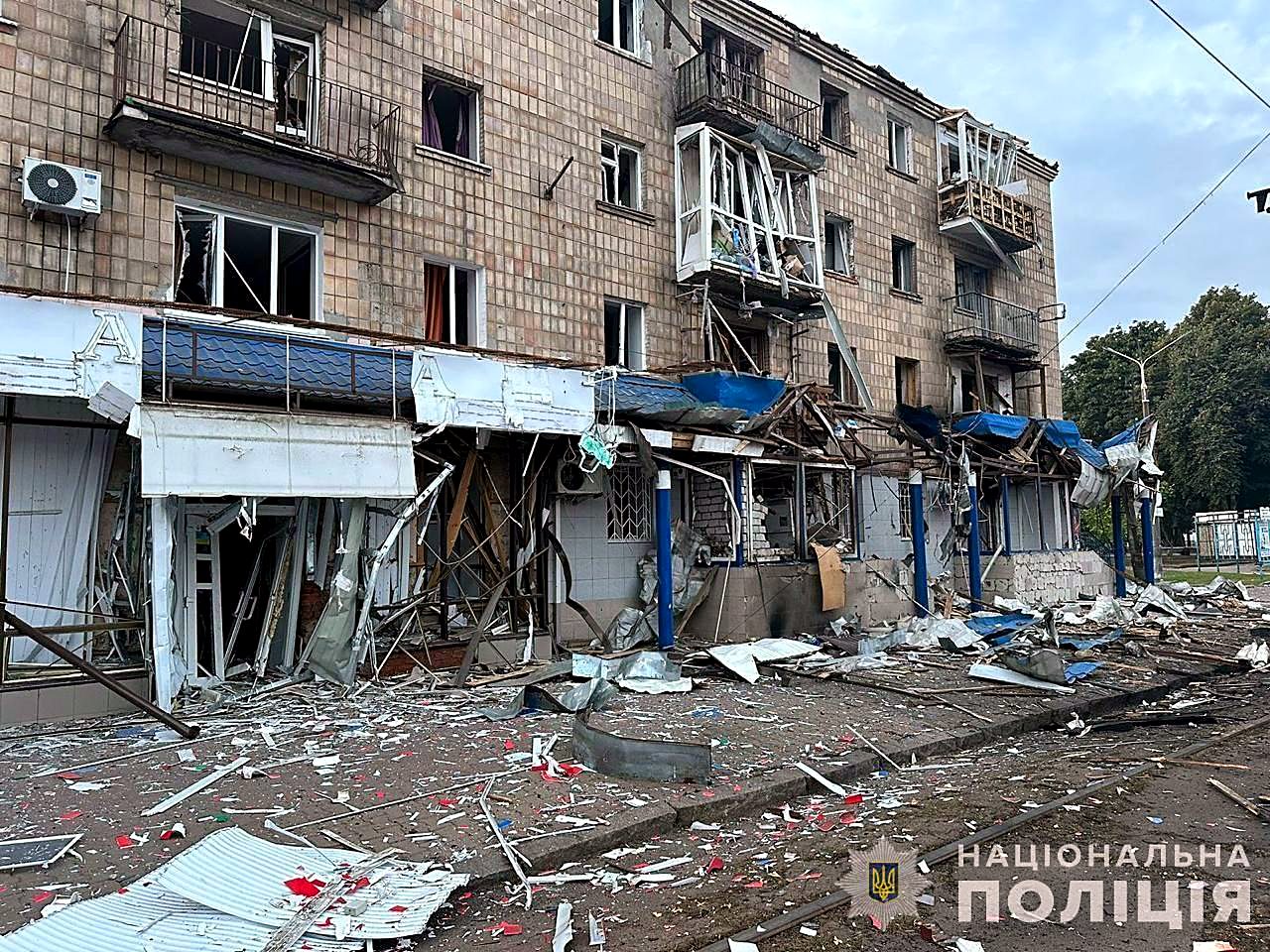 A four-story apartment building with blown-out windows and shattered storefronts, debris covering the street